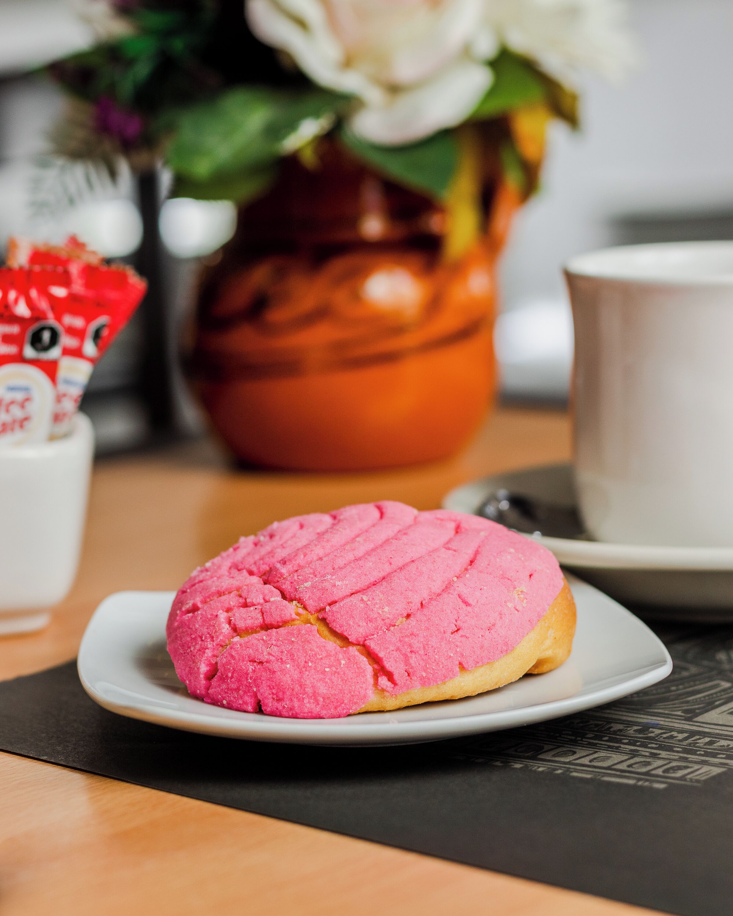 A pink pastry on a white plate, a white cup, and a brown vase with flowers in the background.