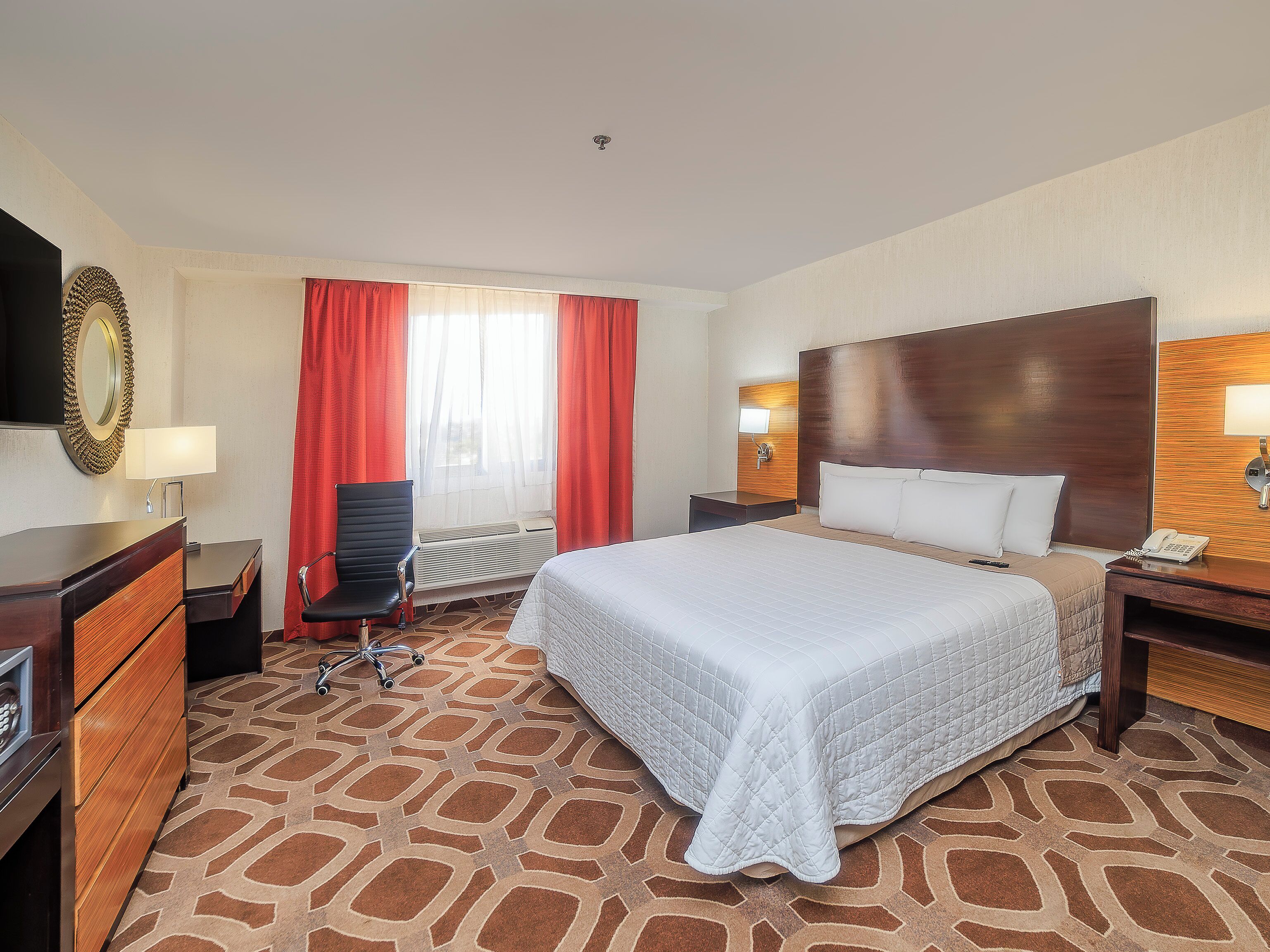 Habitación de hotel con cama, escritorio, silla, espejo, ventana con cortinas y una lámpara de pared.