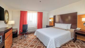 Habitación de hotel con cama, escritorio, silla, espejo, ventana con cortinas y una lámpara de pared.