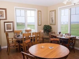 Comedor con una mesa redonda de madera y seis sillas de madera. Hay dos ventanas grandes con marcos blancos que ofrecen vistas al exterior.