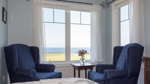 A bedroom with a bed, two blue armchairs, a small table with a vase of flowers, and a view of the beach through large windows. - Johnson Shore Inn (Rock Barra)