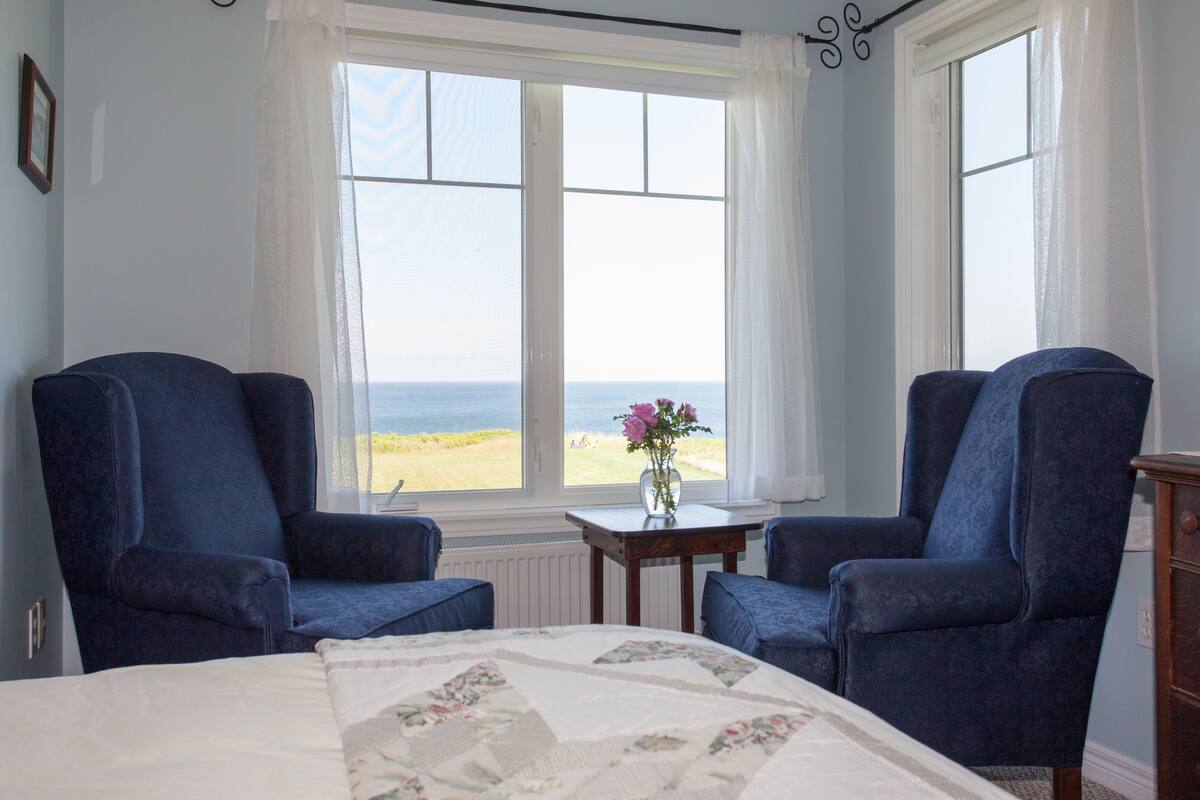 A bedroom with a bed, two blue armchairs, a small table with a vase of flowers, and a view of the beach through large windows.