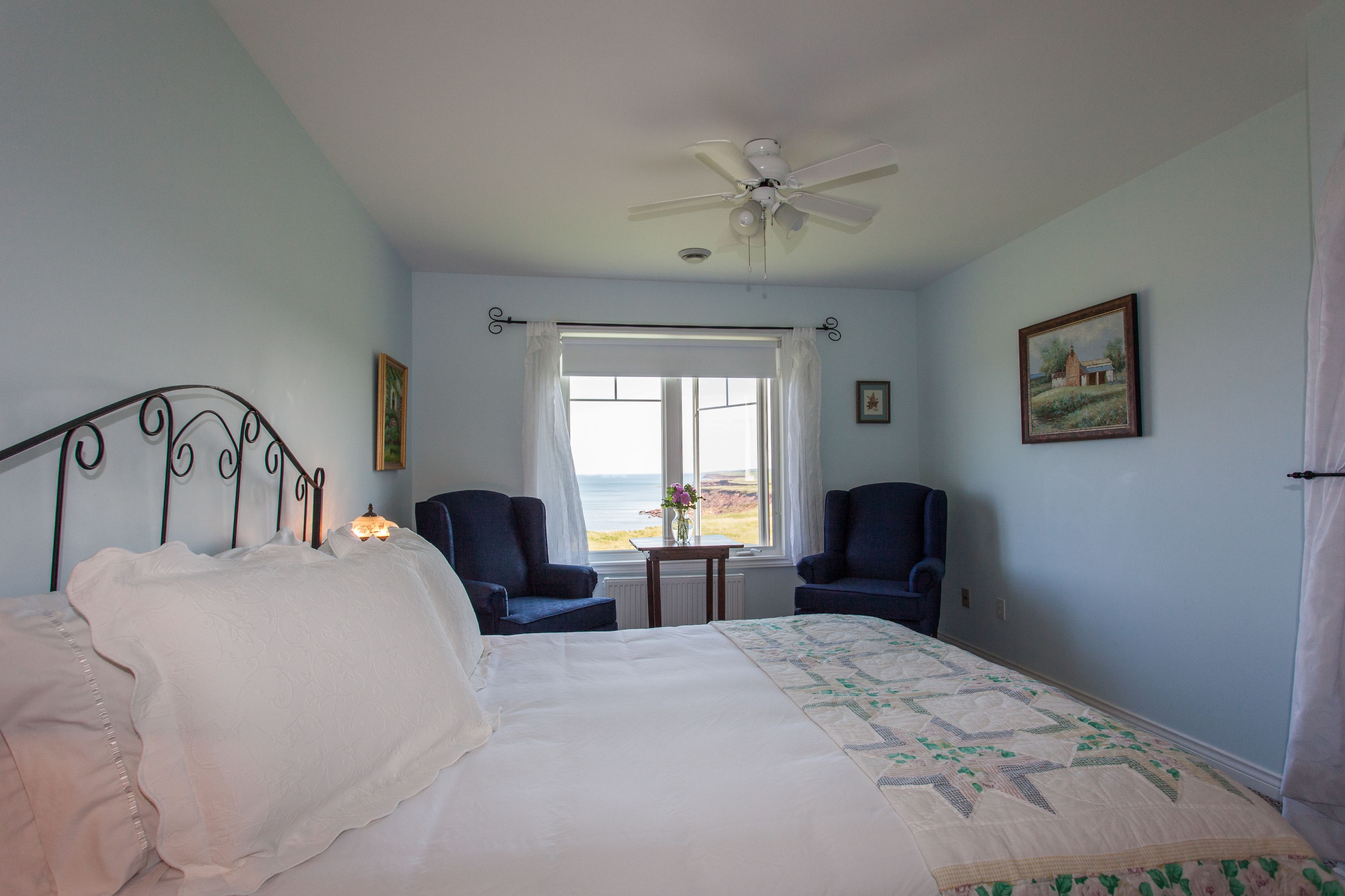 A bedroom with a bed, two chairs, a ceiling fan, a window with a view of the sea, and a painting on the wall.