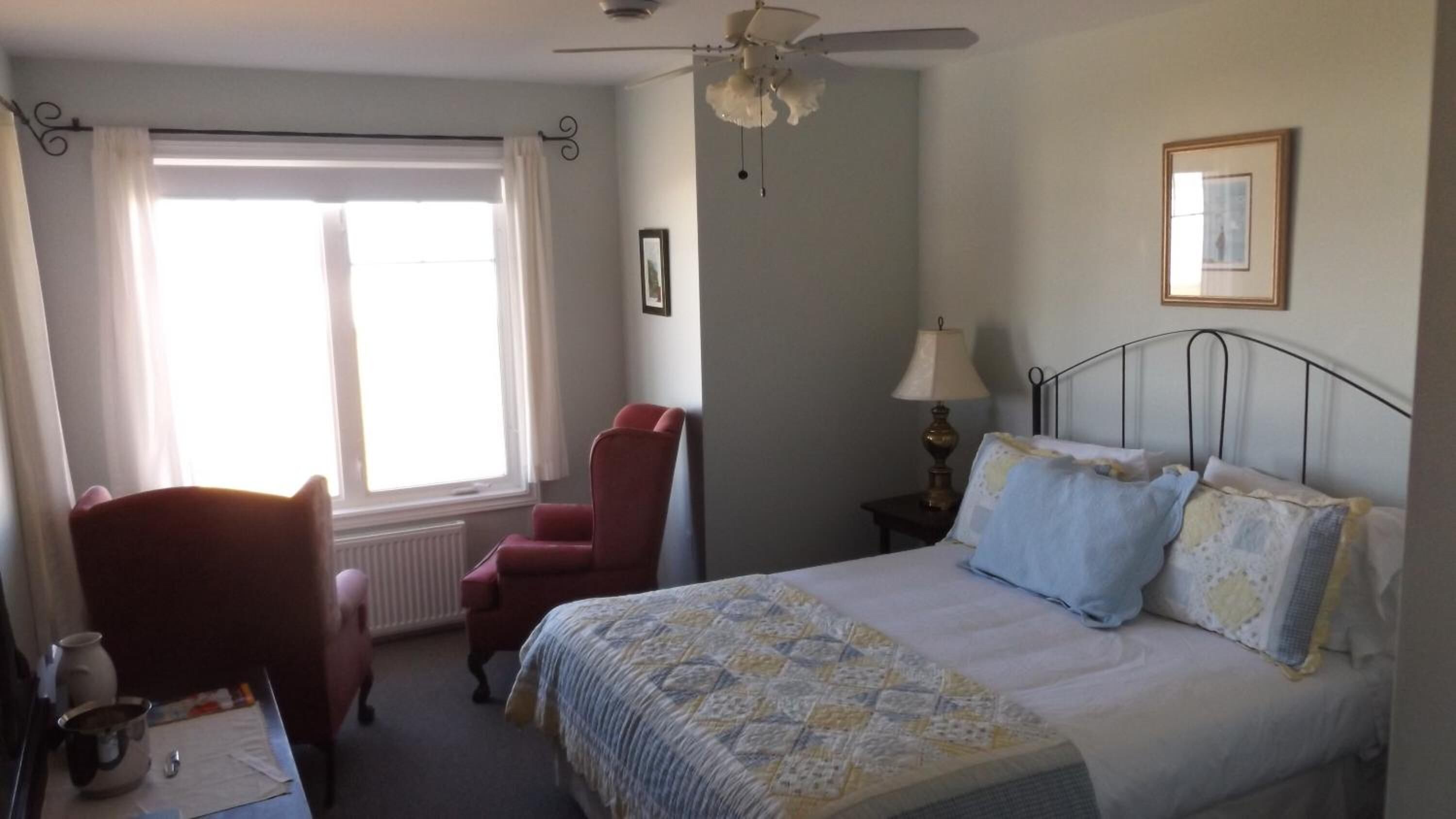 Un dormitorio con cama, una silla, una mesita de noche, lámpara, ventana con cortinas y ventilador de techo.