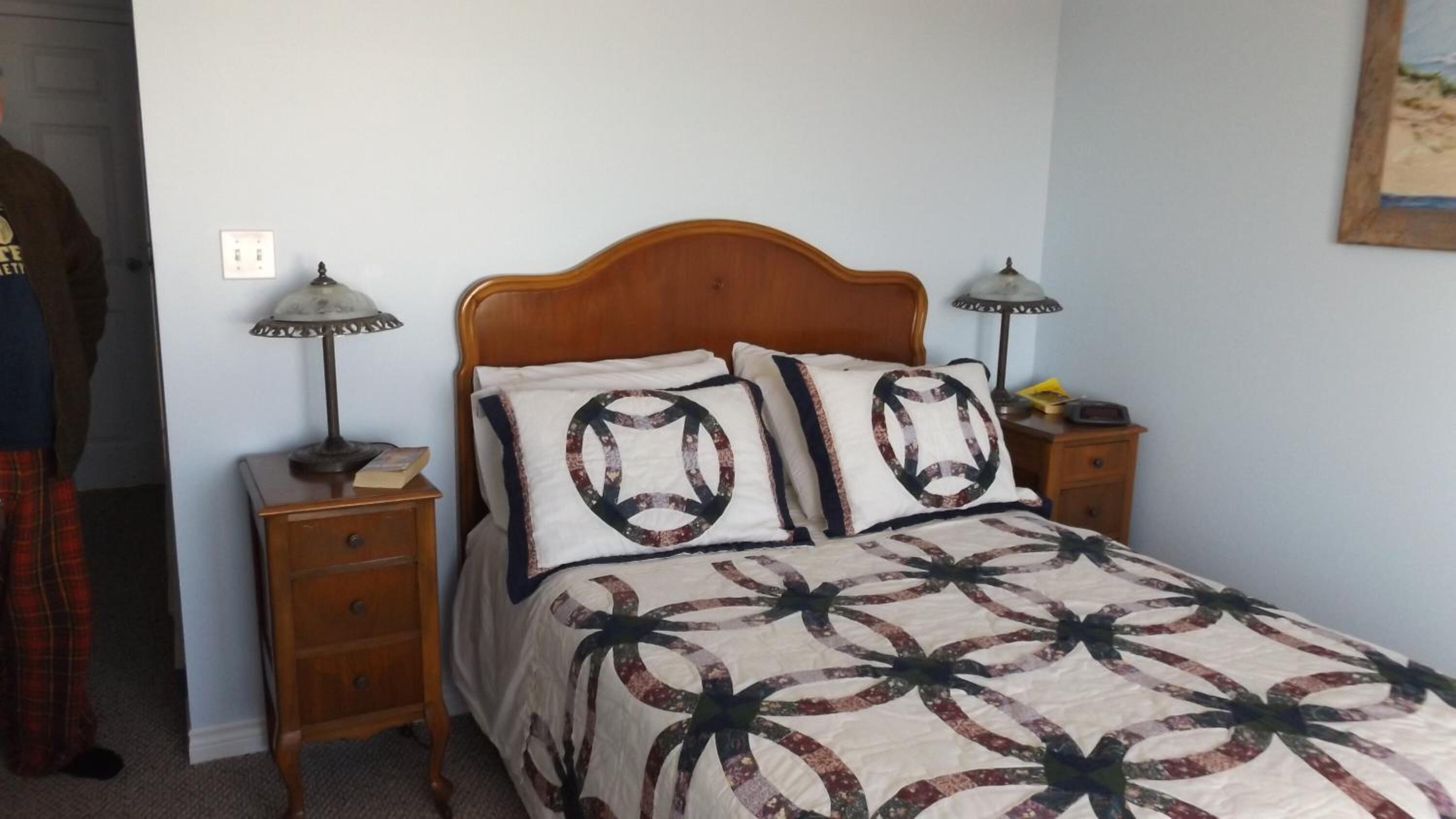Un dormitorio con cabecera de madera, dos mesitas de noche con lámparas, una cama con colcha estampada y una mesita auxiliar con un libro y una lámpara.