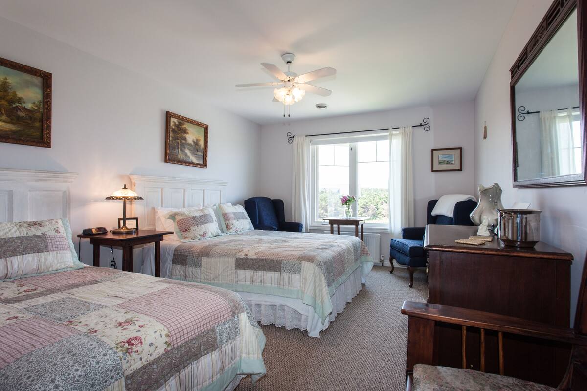 A bedroom with two beds, a ceiling fan, a window with curtains, a chair, a table with a lamp, and a mirror.