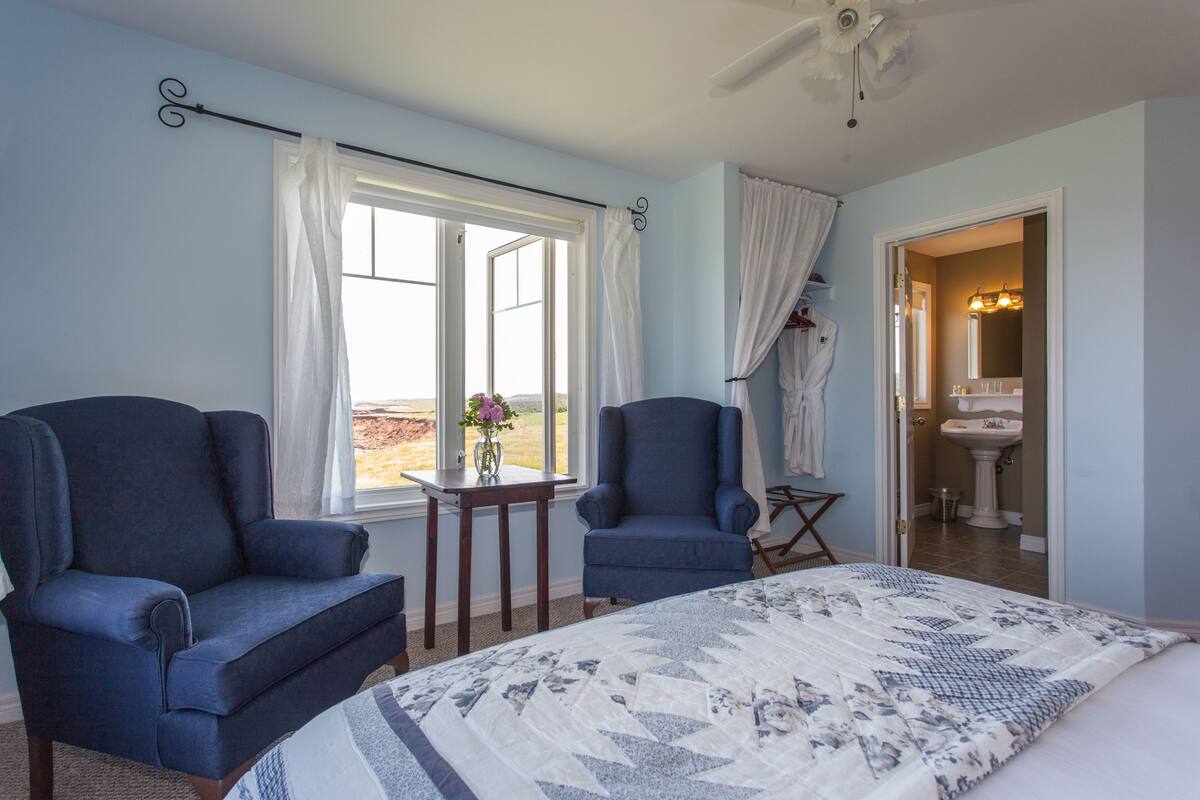 A bedroom with two armchairs, a bed with a patterned quilt, a window with curtains, and a bathroom visible through an open door.