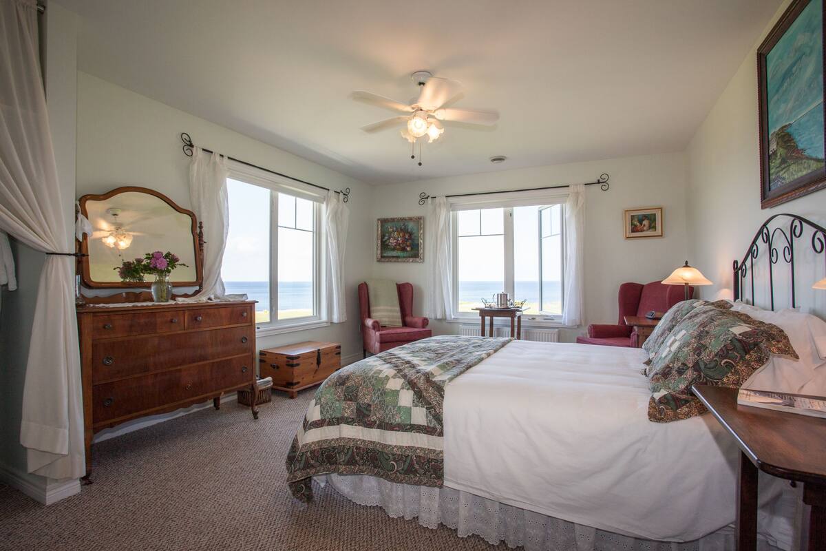 A bedroom with a large bed, a dresser, a chair, and a desk with a lamp. There are windows with curtains, a ceiling fan, and a painting on the wall.