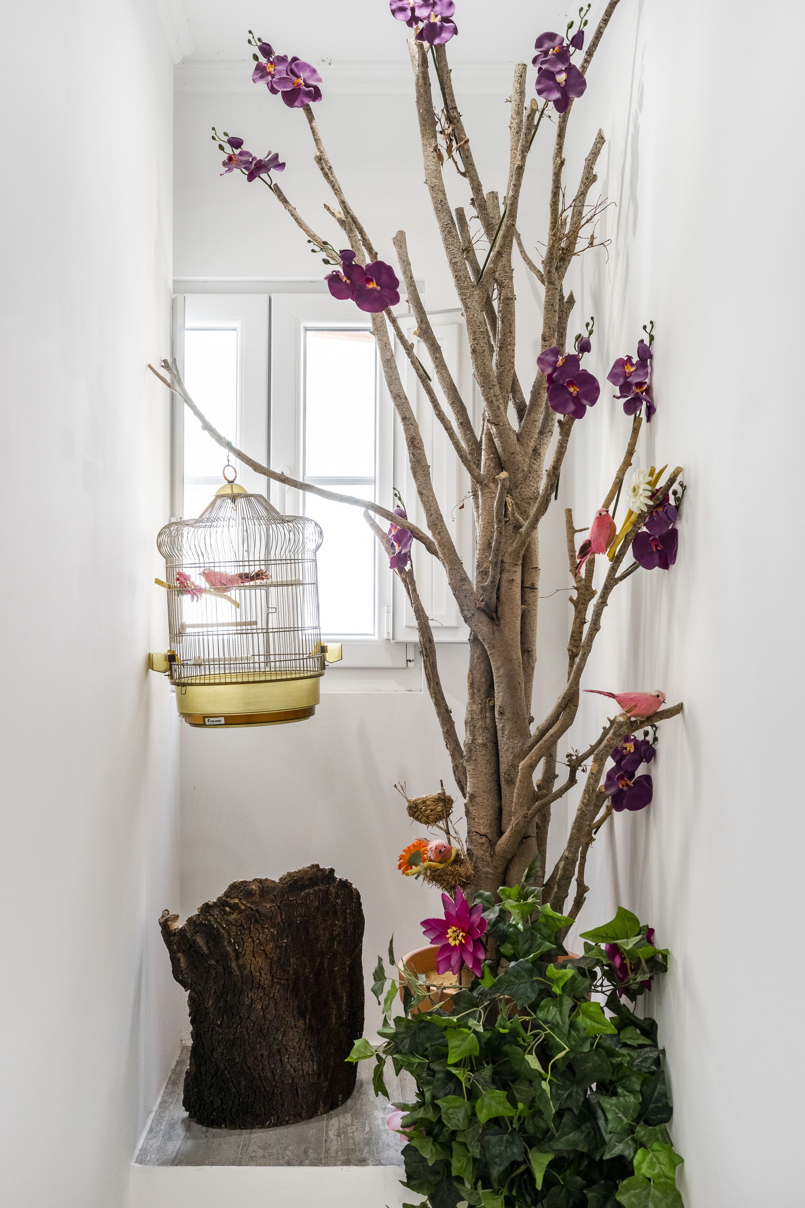 A room with a wooden tree sculpture, a hanging birdcage, and a potted plant with flowers.