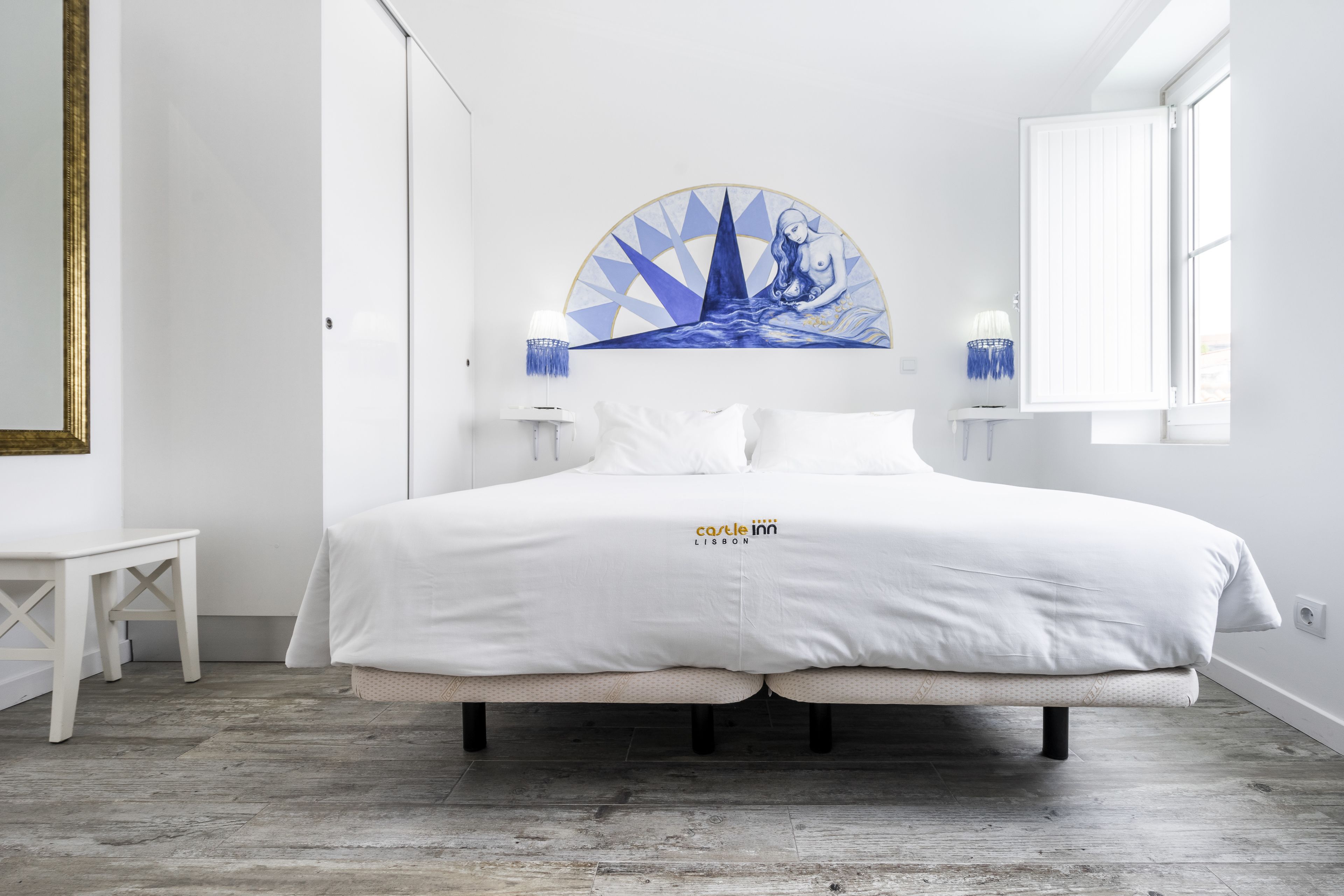 Une chambre à coucher avec un grand lit, une table de chevet blanche, un tableau accroché au mur et une fenêtre avec des rideaux.