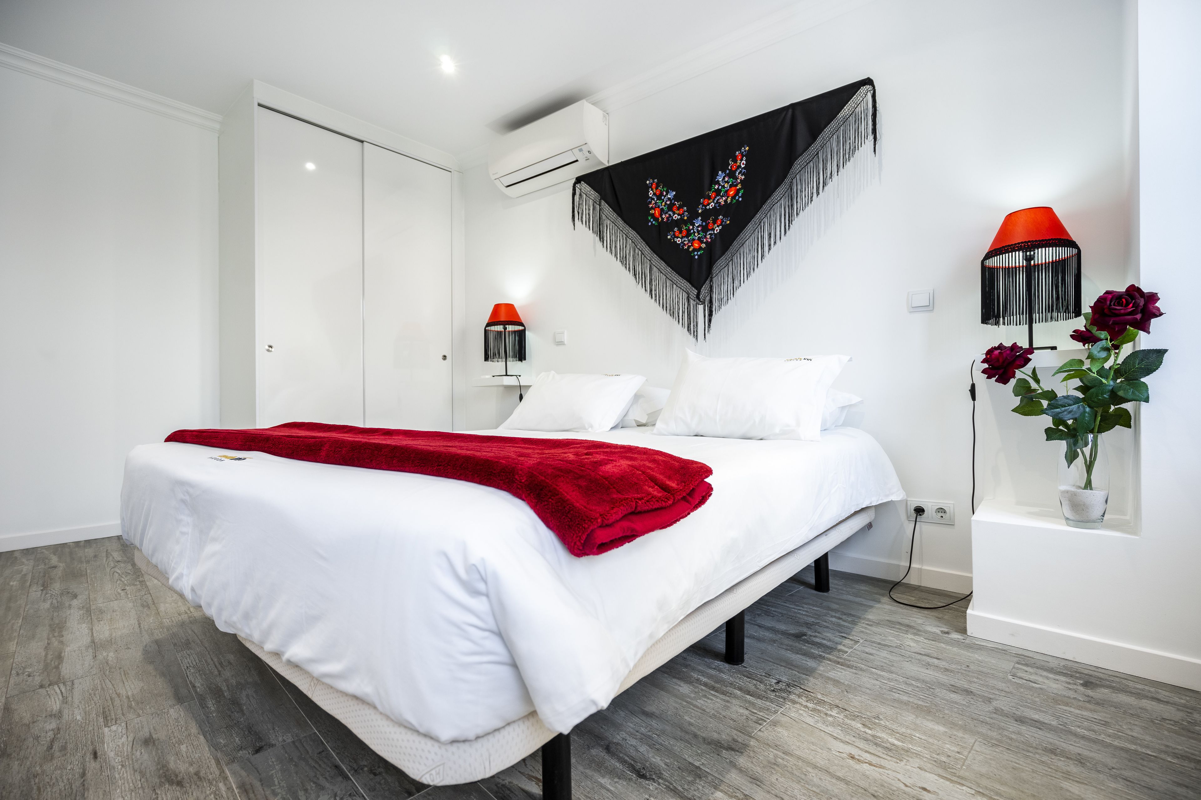 A modern bedroom with a large bed, white bedding, a red throw blanket, a black and white tapestry, a red lamp, and a potted plant with red roses.