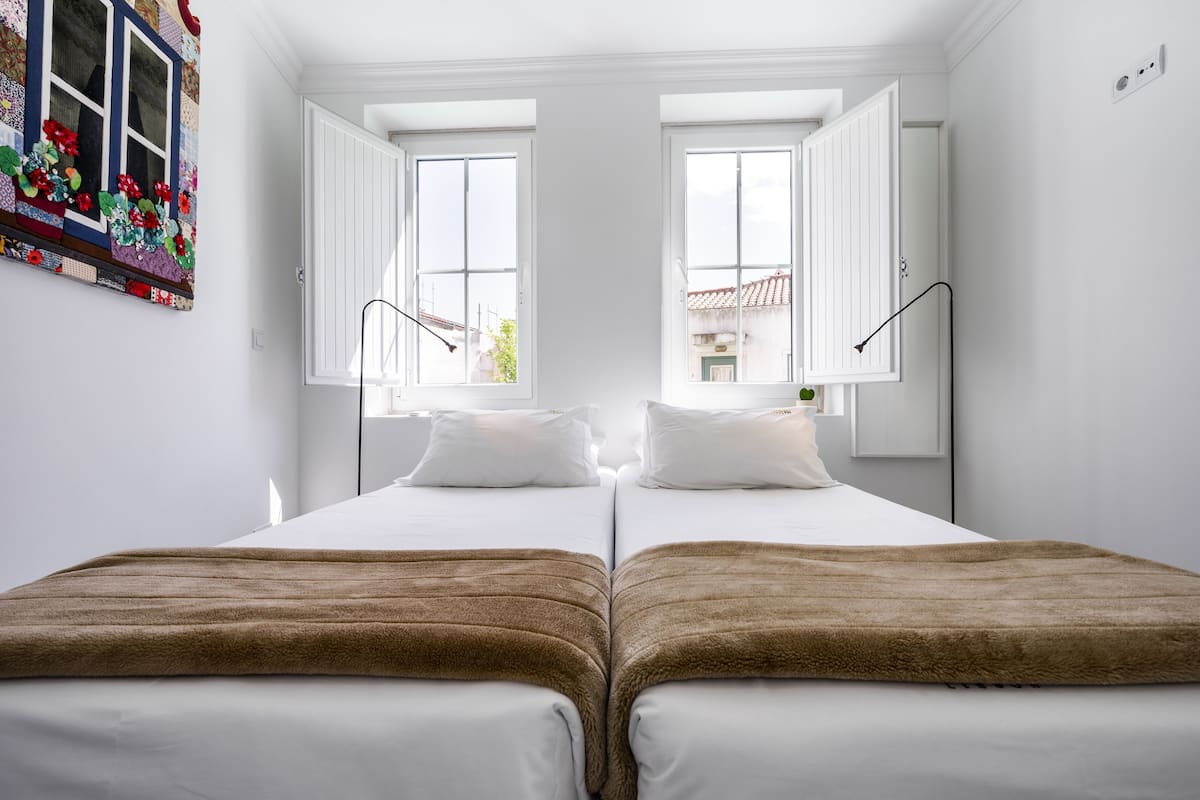 Una cama doble con sábanas blancas y una manta marrón, dos ventanas con contraventanas abiertas y una lámpara fijada a la pared.