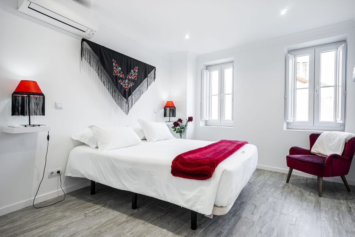 Un dormitorio moderno con una cama grande, dos lámparas de noche, una silla roja y una ventana con cortinas blancas.