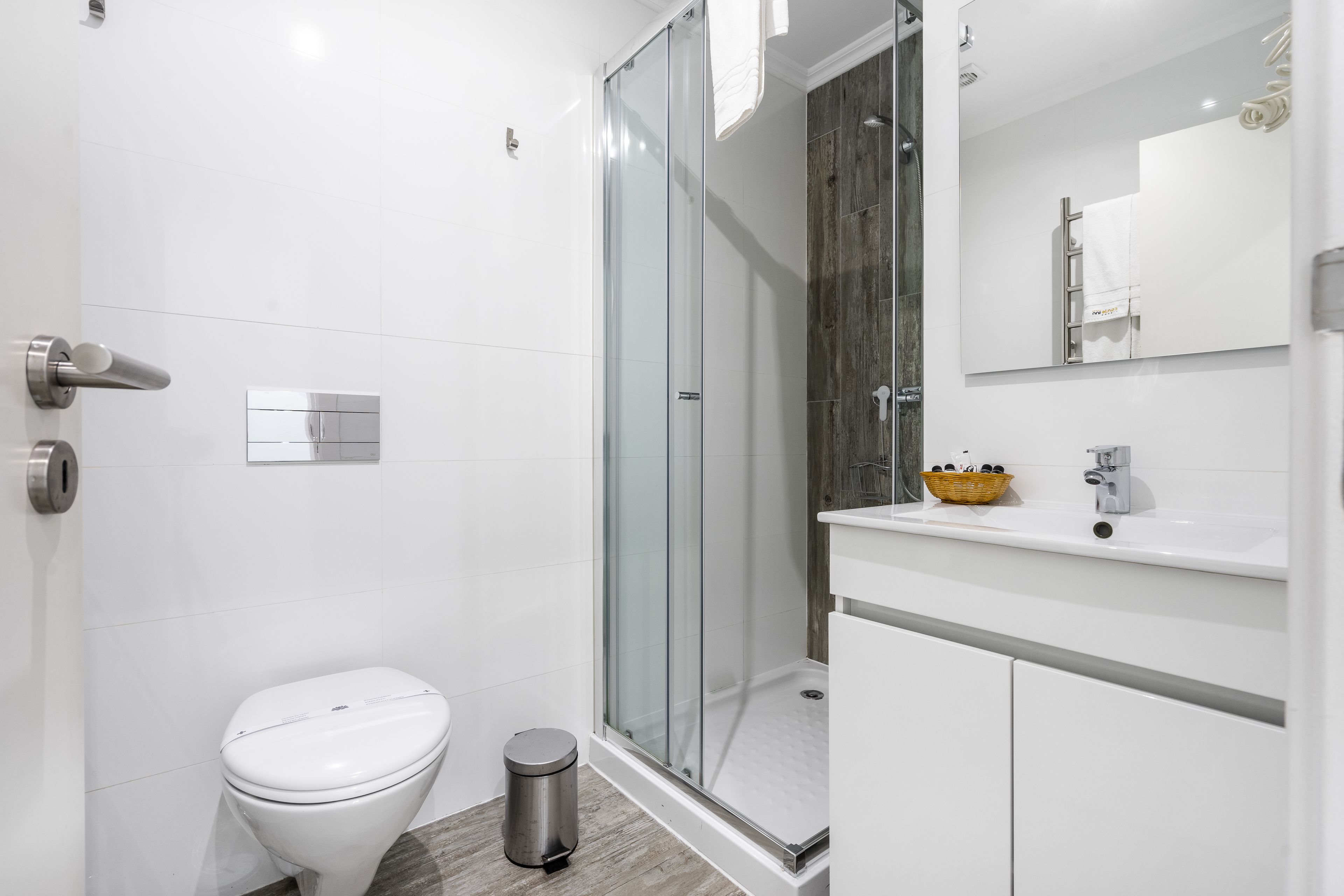 Un baño moderno con inodoro, ducha con cerramiento de vidrio, lavamanos con espejo y un pequeño cubo de basura.