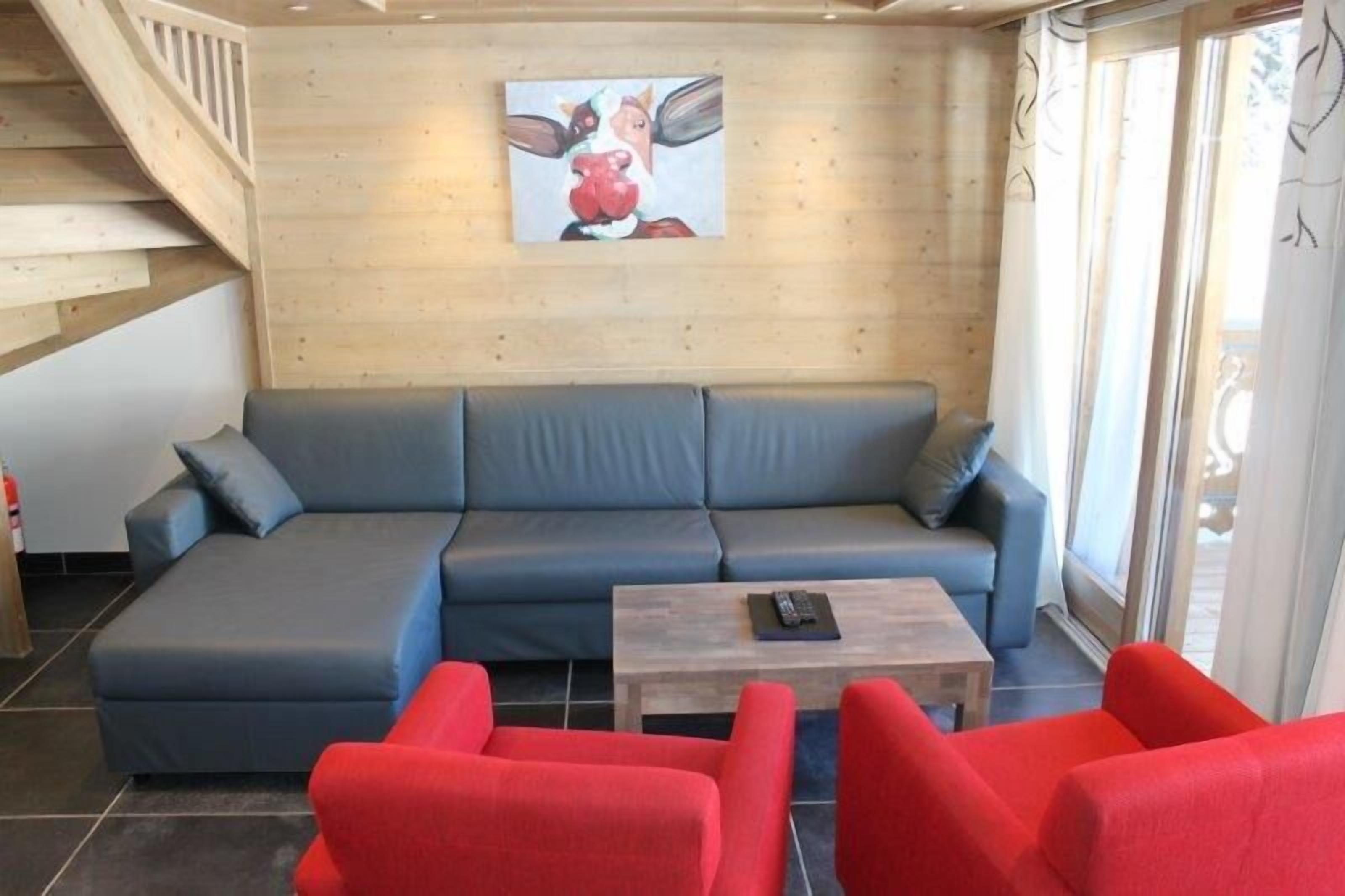 A modern living room with a grey sofa, red armchairs, a wooden coffee table, and a painting of a cow on the wall.