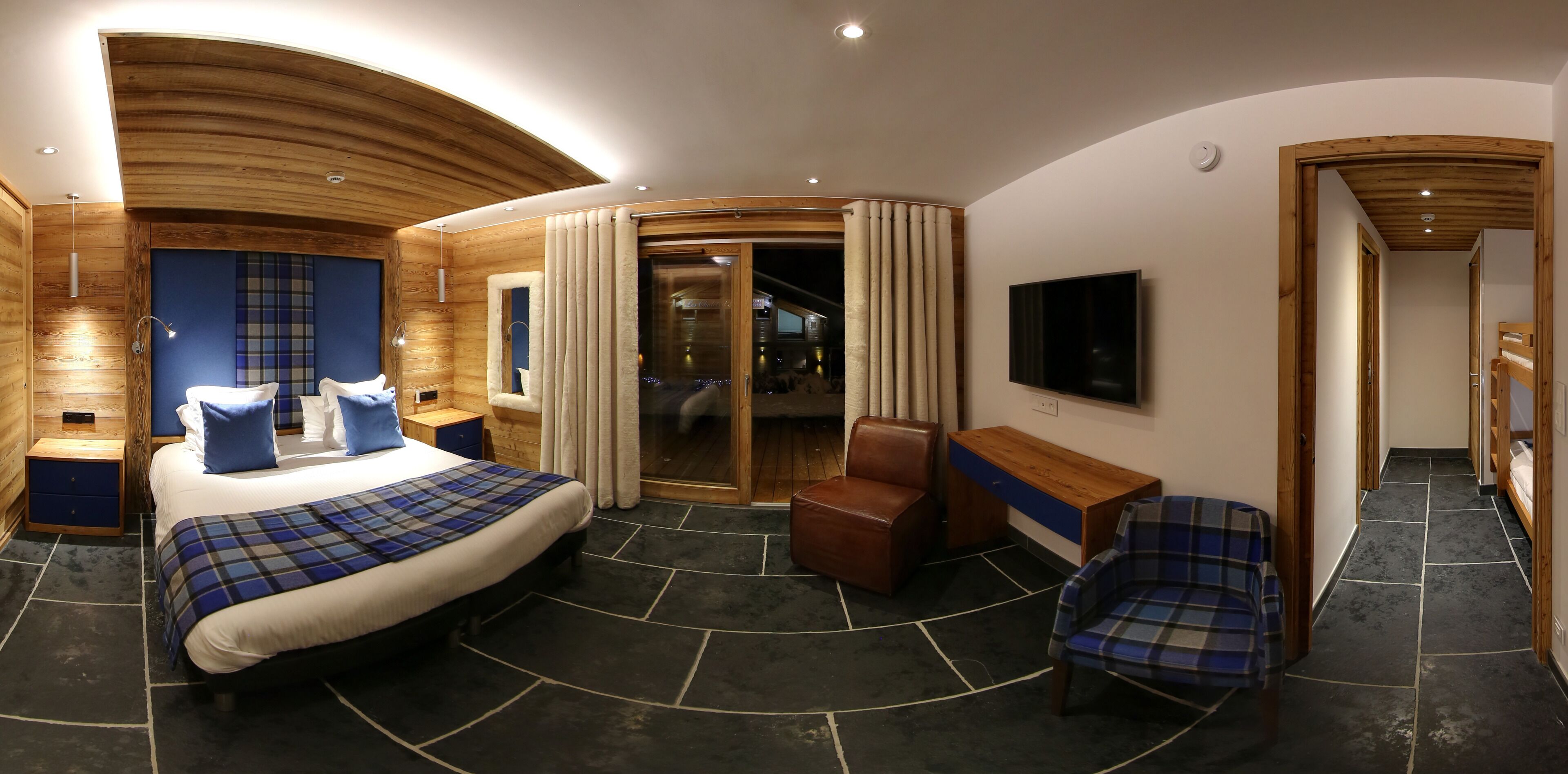 A modern hotel room with a large bed, a wooden ceiling, a TV, a seating area, and a sliding glass door leading to a balcony.