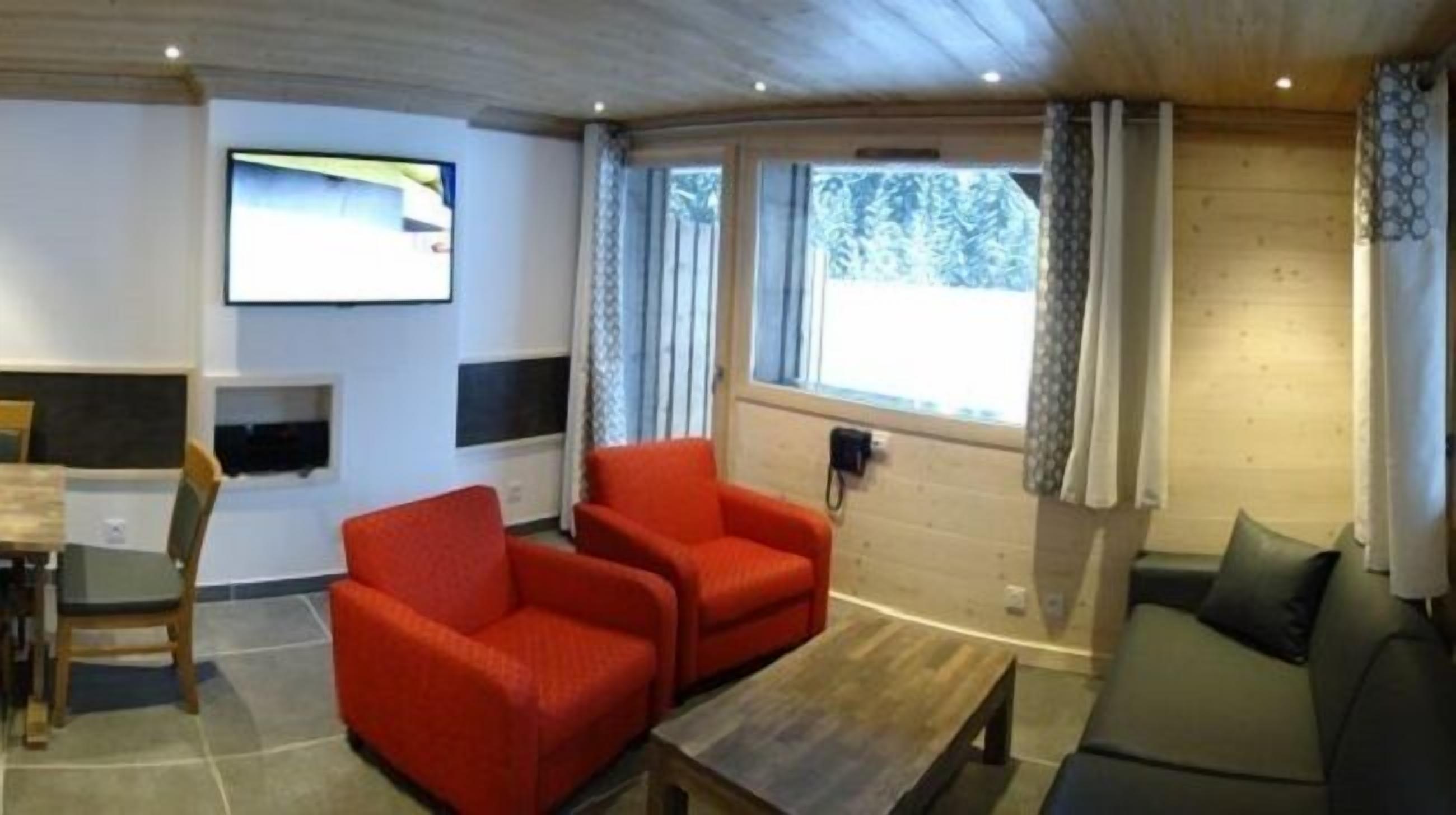 A room with two red armchairs, a sofa, a wooden coffee table, a TV mounted on the wall, and a window with curtains.