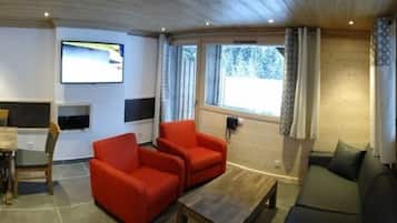A room with two red armchairs, a sofa, a wooden coffee table, a TV mounted on the wall, and a window with curtains.
