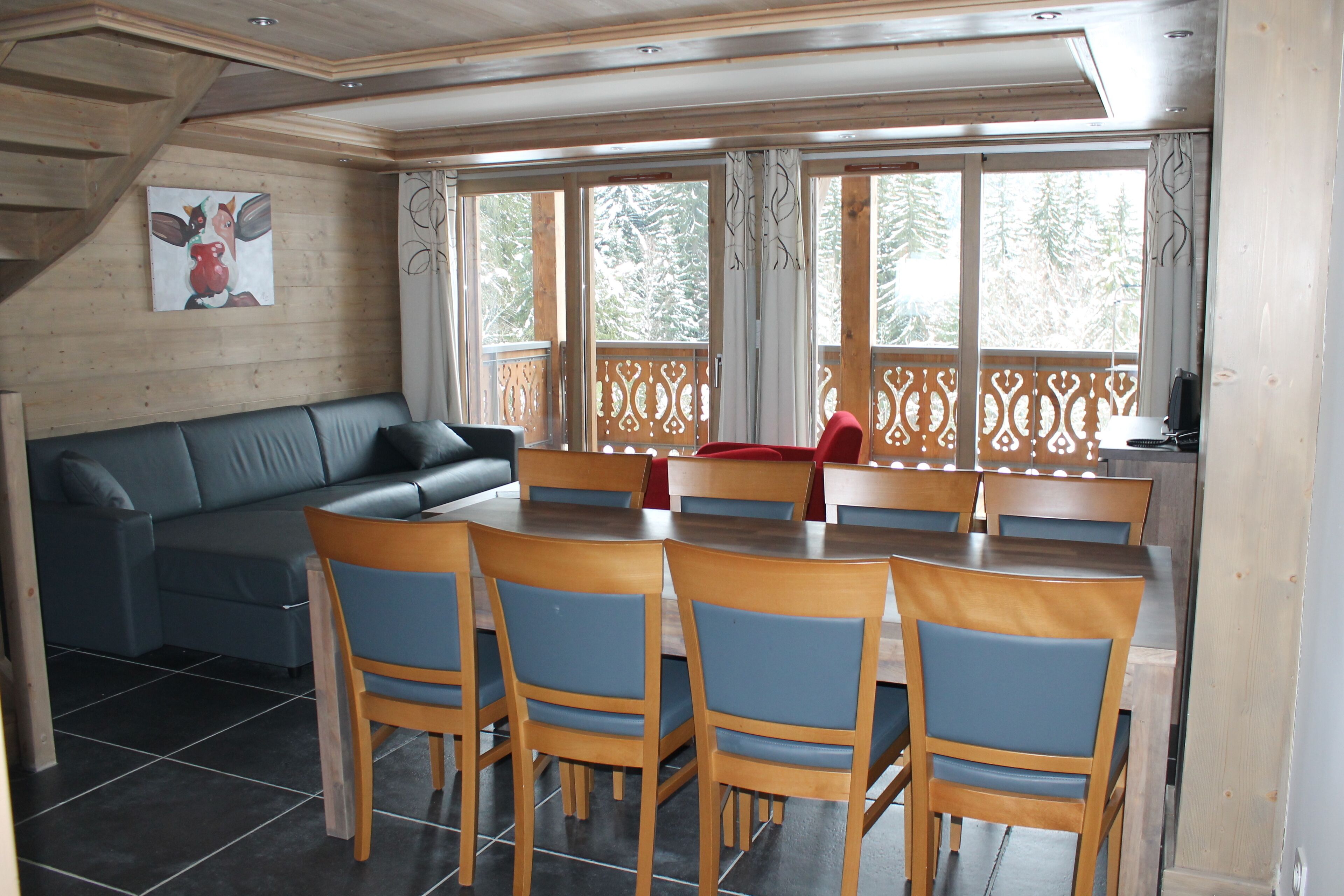 A dining area with a wooden table and chairs, a sofa, and a view through large windows.