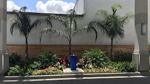 Un edificio con fachada de ladrillo blanco, terraza de madera y jardín ajardinado con palmeras y flores coloridas.