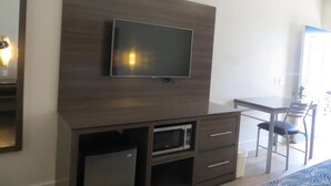 Una habitación moderna con televisión de pantalla plana montada en un panel de madera oscura, un microondas en un mueble debajo, una zona de comedor con mesa y sillas, y un espejo grande a la izquierda.