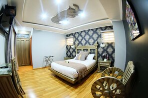Un dormitorio con cama, piso de madera, ventilador de techo, ventana con persianas y televisor montado en la pared.