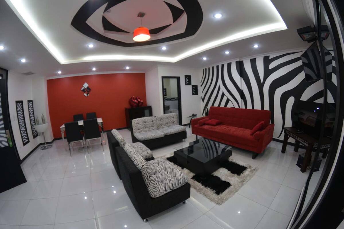 A modern living room with a red sofa, black and white patterned sofas, a glass coffee table, and a zebra-patterned wall.