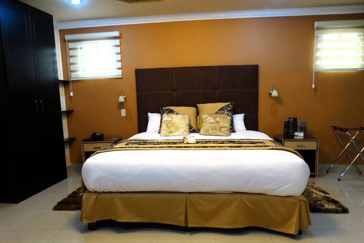 A hotel room with a large bed, a nightstand, a lamp, and a window with blinds.