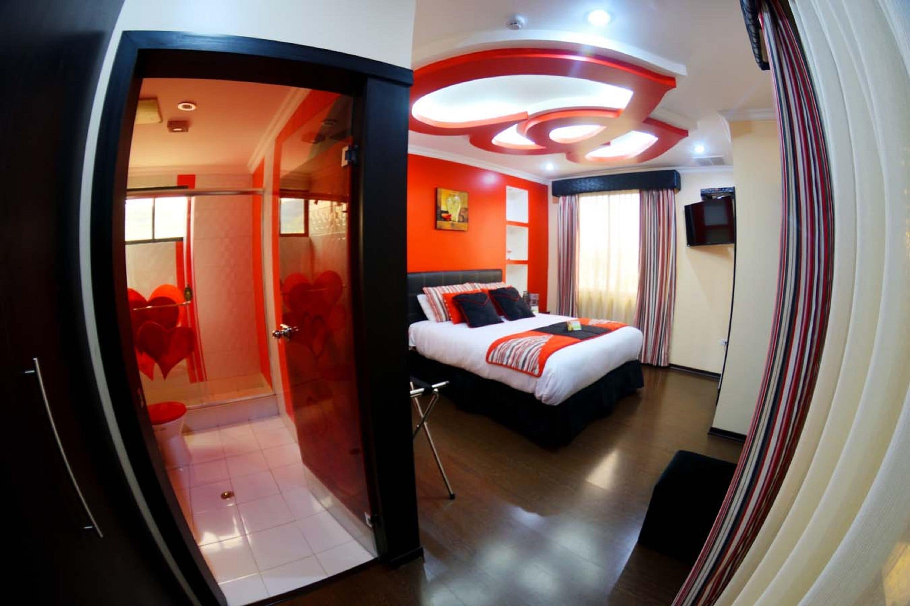 Una habitación de hotel moderna con techo curvo, una cama grande con edredón rojo y blanco, un televisor y zona de ducha.