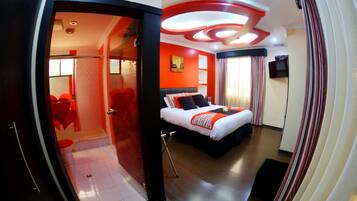 Una habitación de hotel moderna con techo curvo, una cama grande con edredón rojo y blanco, un televisor y zona de ducha.
