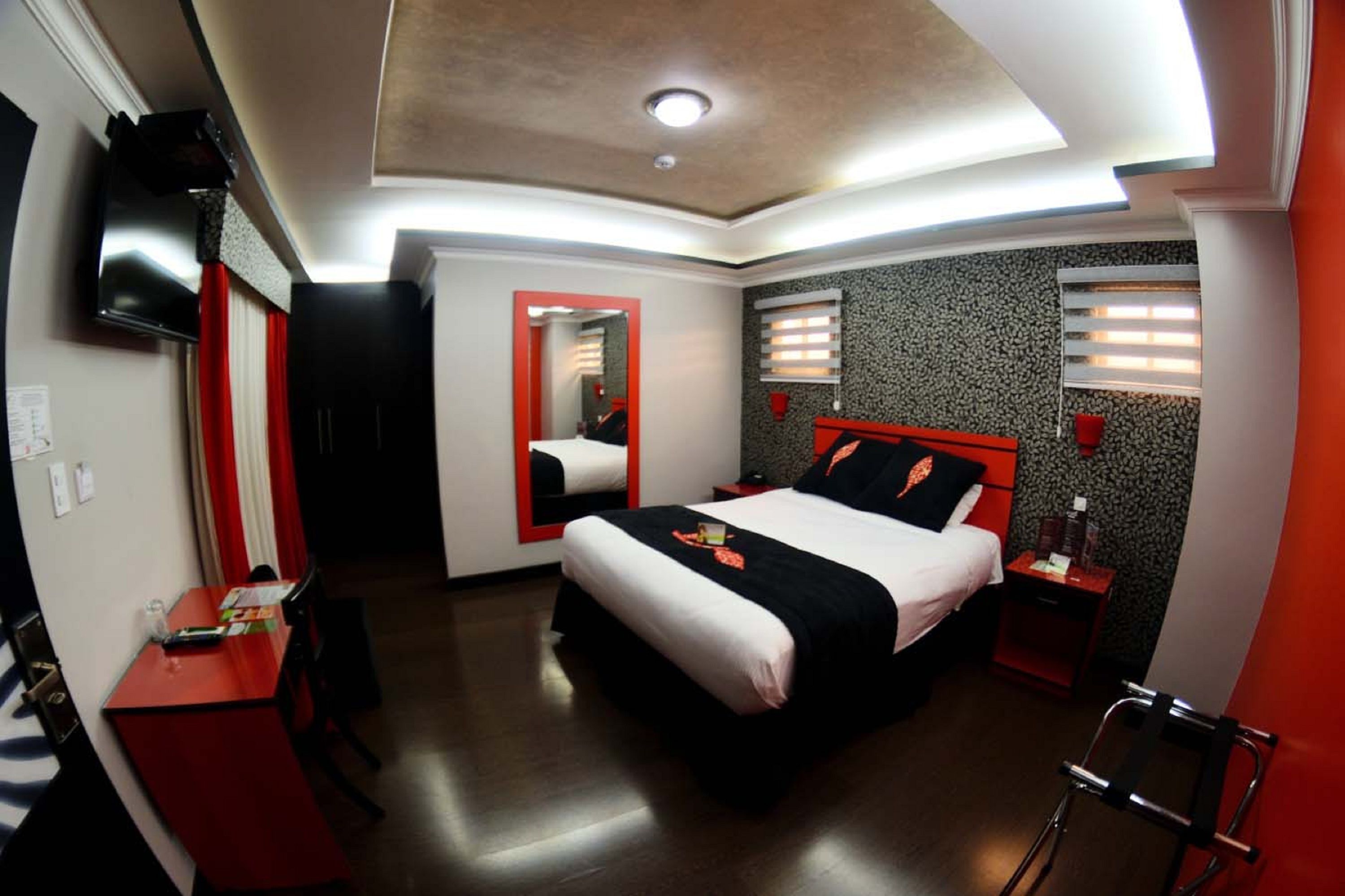 A modern hotel room with a large bed, a desk, a chair, a TV, and a bathroom with a mirror and red accents.
