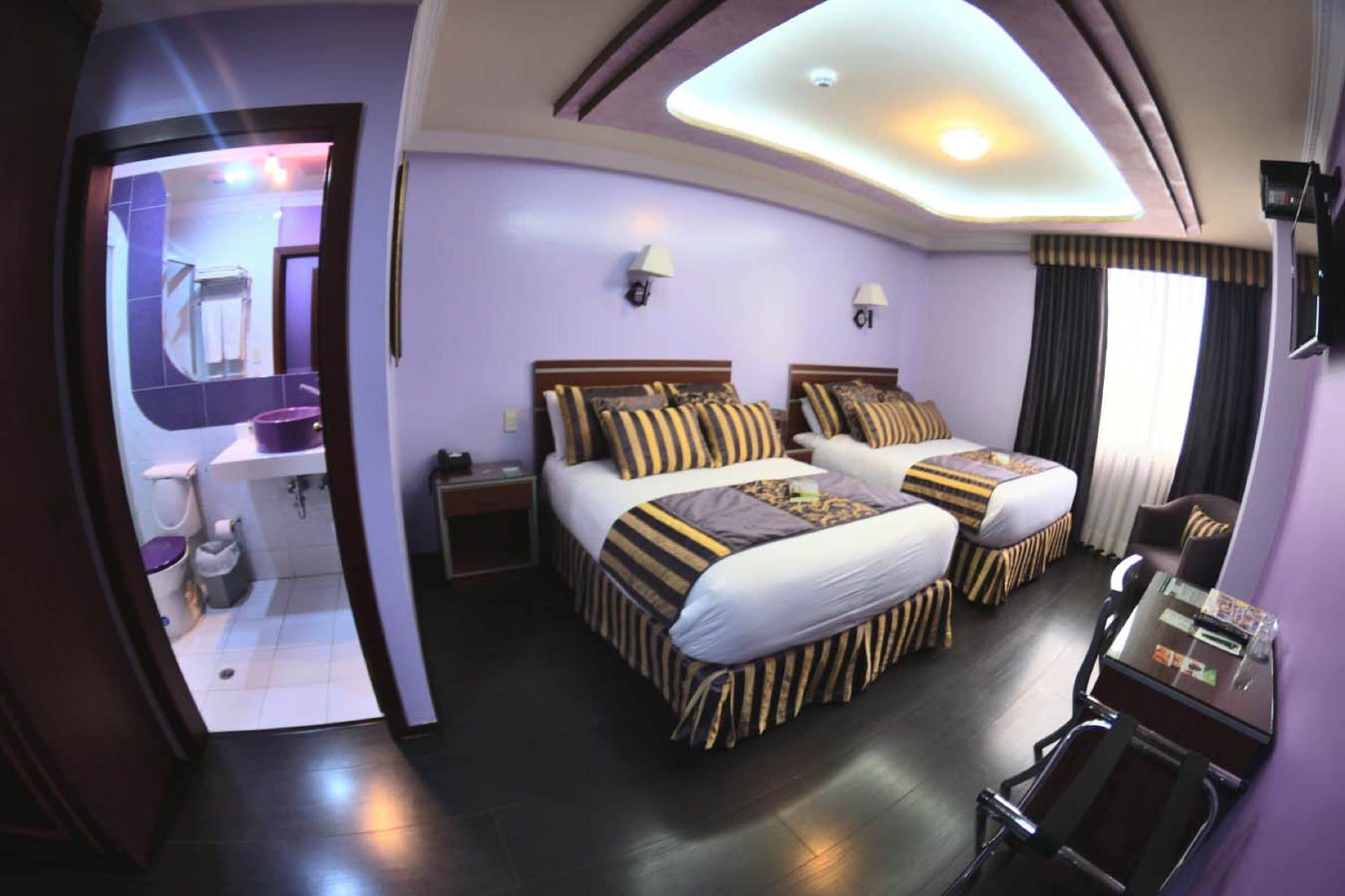 Habitación de hotel con dos camas, un escritorio, una silla, un espejo y un baño visible a través de una puerta abierta.