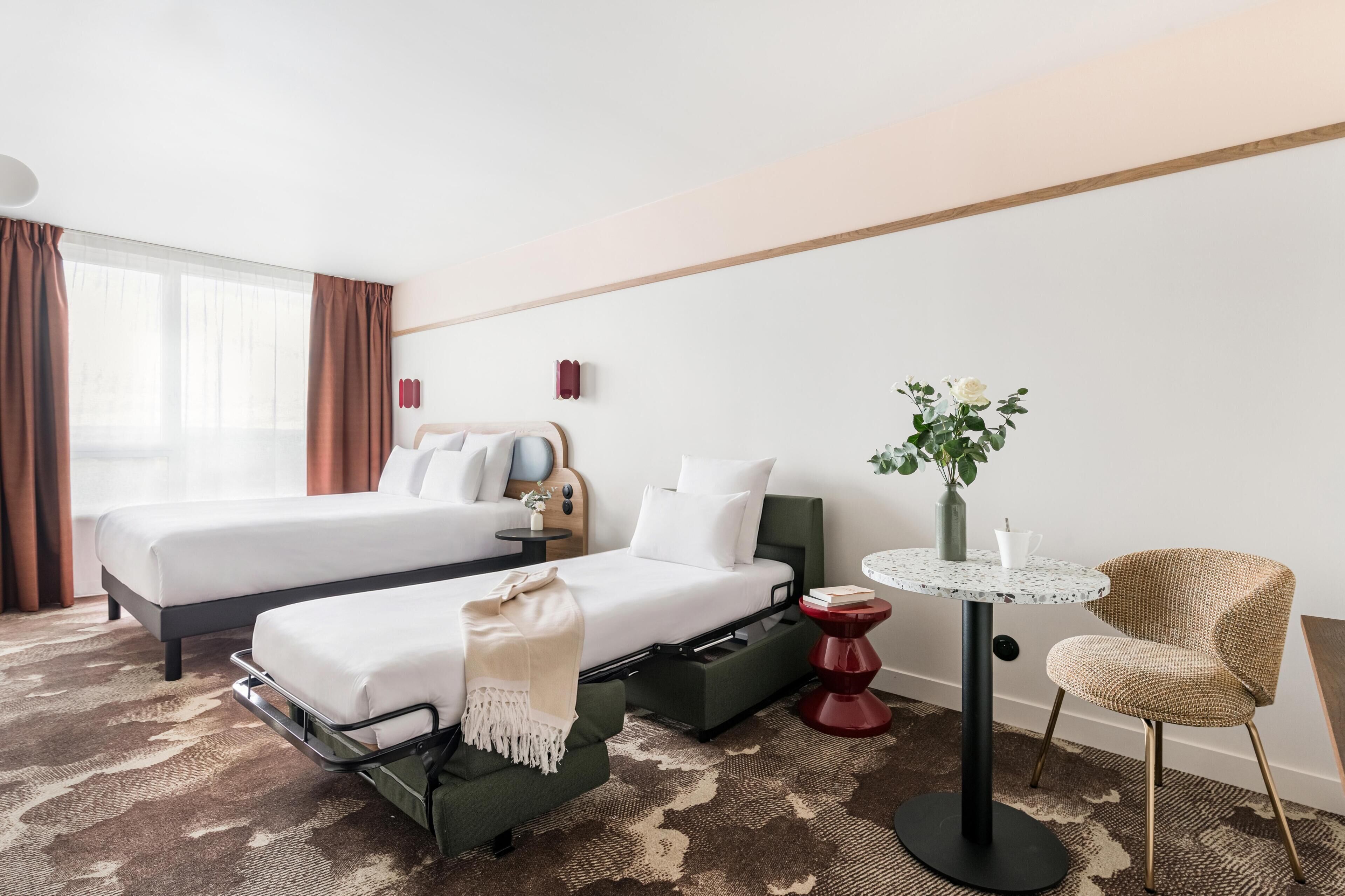 A hotel room with two beds, a small table, a chair, a vase with flowers, and a window with curtains.