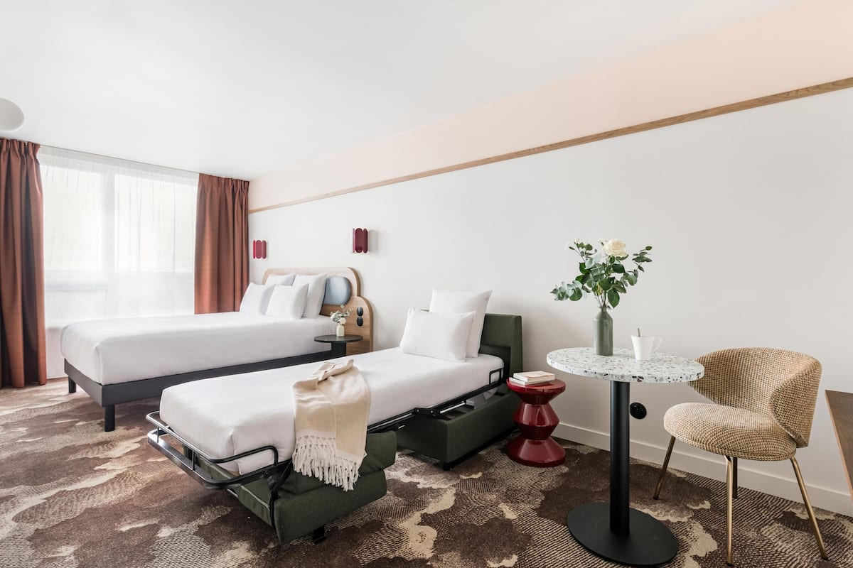 Une chambre d’hôtel avec deux lits, une petite table, une chaise, un vase avec des fleurs et une fenêtre avec des rideaux.