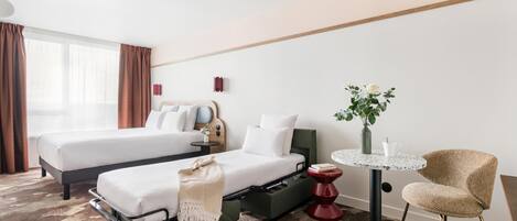 Habitación de hotel con dos camas, una mesita, una silla, un florero con flores y una ventana con cortinas.