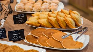A buffet table with various food items including pancakes, pastries, and a sign indicating a madeleine.