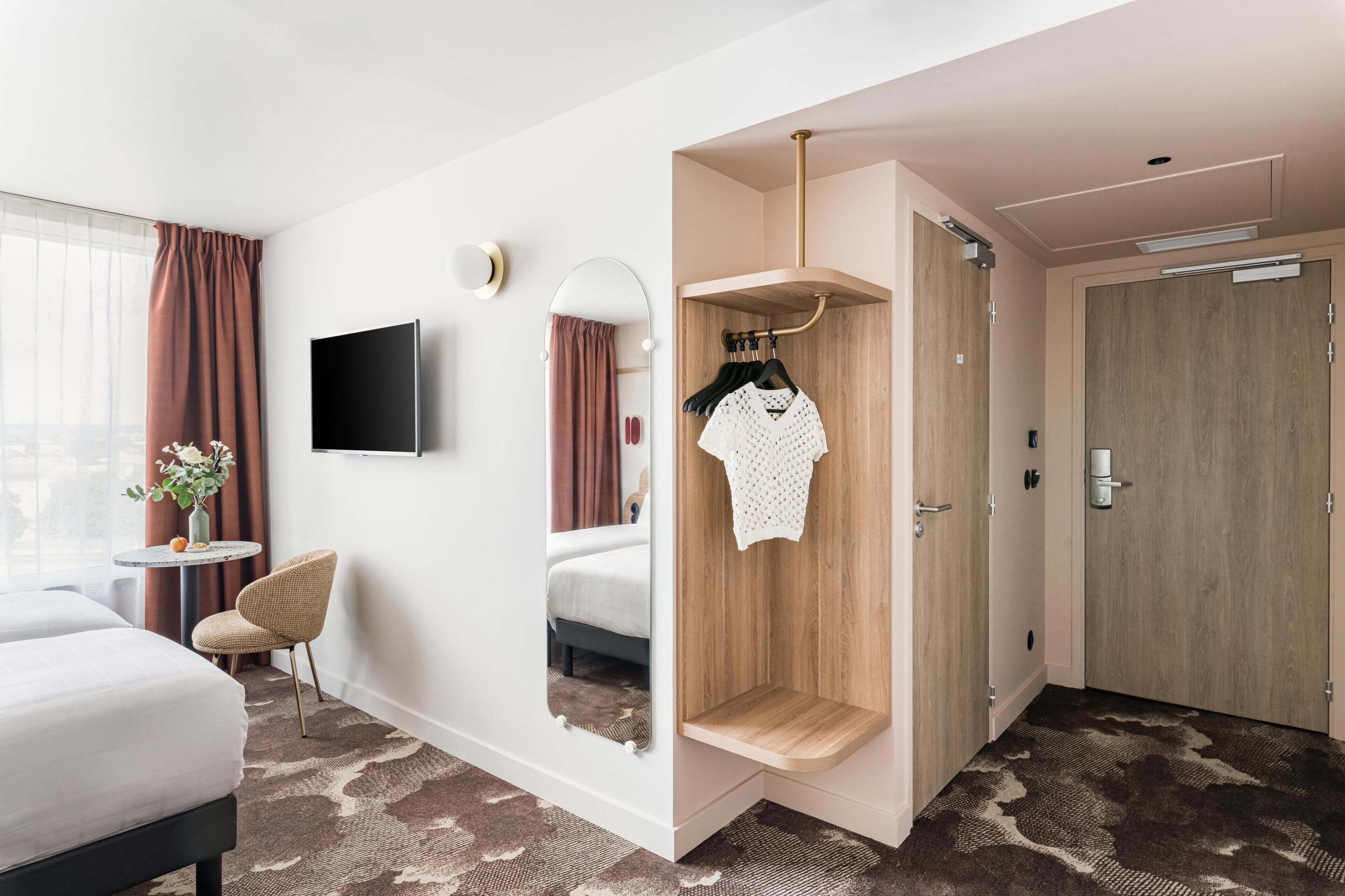 Ein Hotelzimmer mit Bett, Fernseher, Spiegel, einem kleinen Tisch mit einer Vase und einem Kleiderschrank.