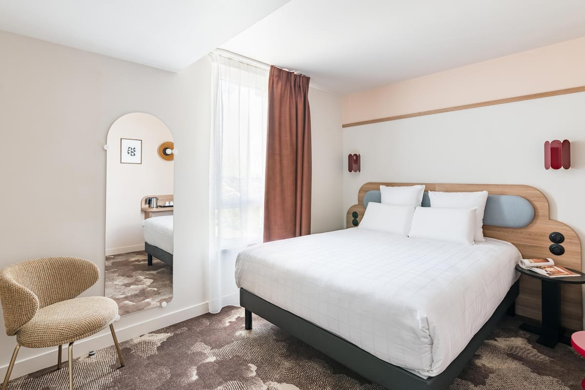 Une chambre d’hôtel avec un lit, une chaise, une table de chevet, un miroir et une fenêtre avec des rideaux.