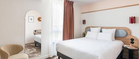 Habitación de hotel con cama, sillón, mesita de noche, espejo y ventana con cortinas.