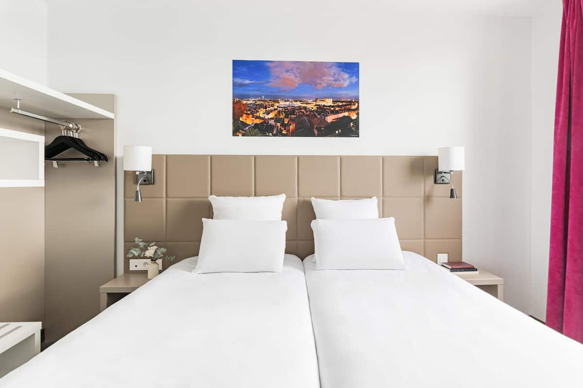 Une chambre d’hôtel avec deux lits, une vue sur la ville, une tête de lit, des lampes de chevet, une table de chevet et un porte-manteau mural.