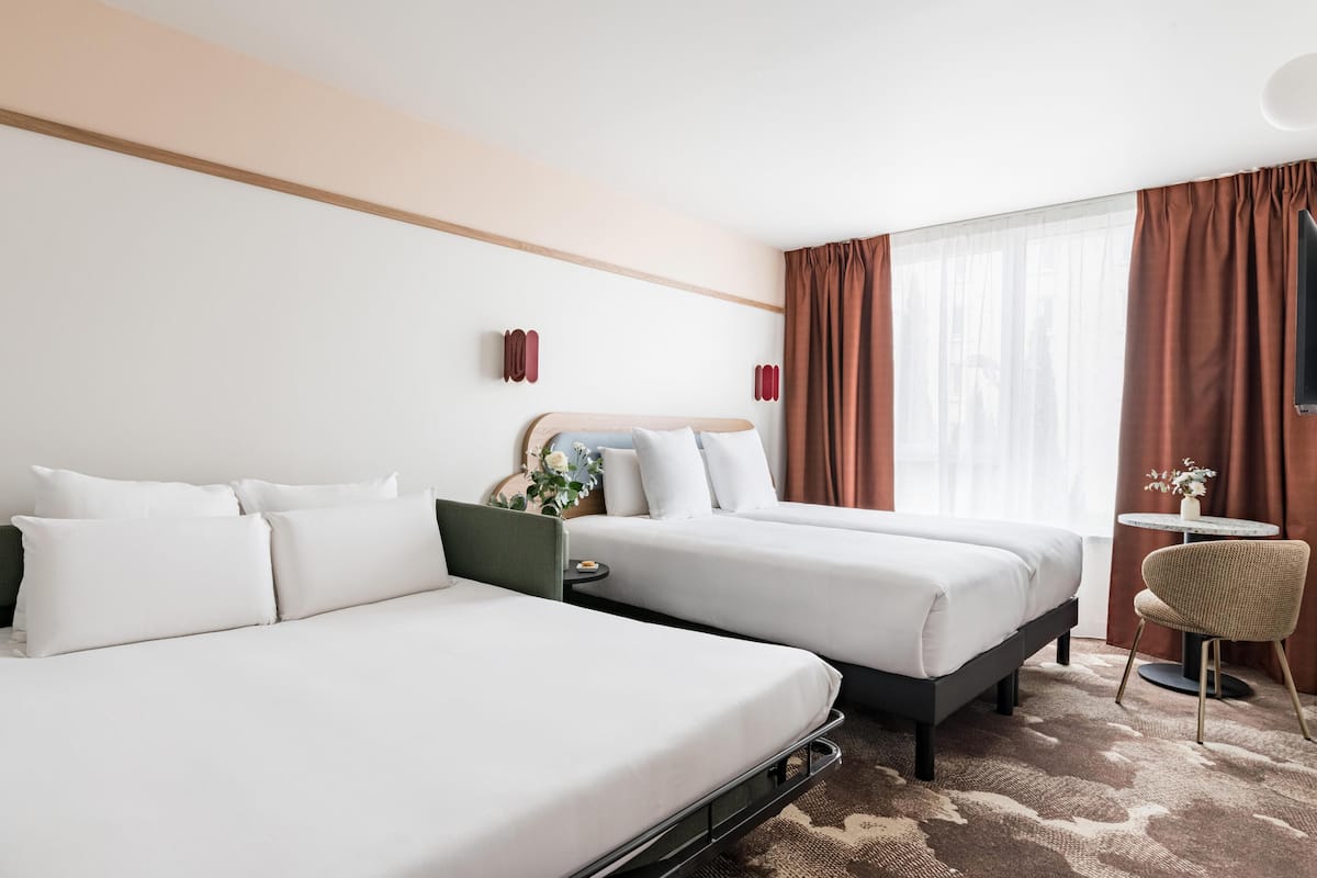 Une chambre d’hôtel avec deux lits, une petite table, une chaise et une fenêtre avec des rideaux.