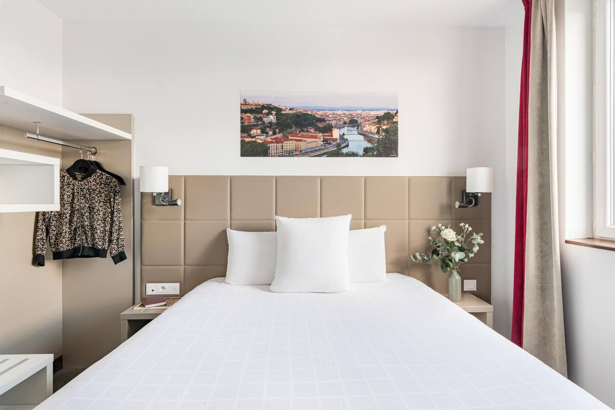 Une chambre d’hôtel avec un lit, une vue sur la ville, un rideau rouge et un vase rempli de fleurs.