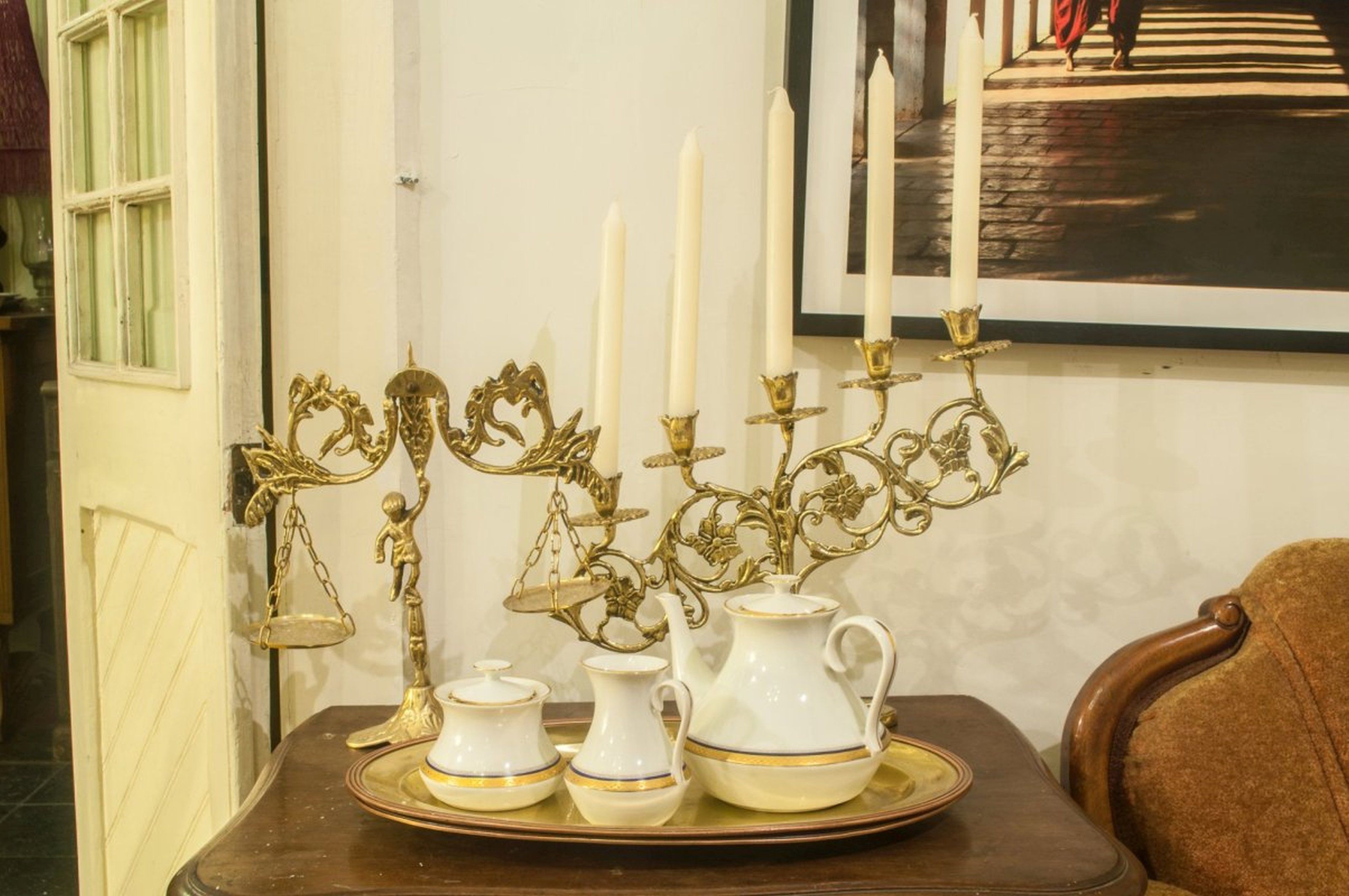 A table with a white pitcher, a teapot, and a sugar bowl on a tray, accompanied by a candelabra with candles.