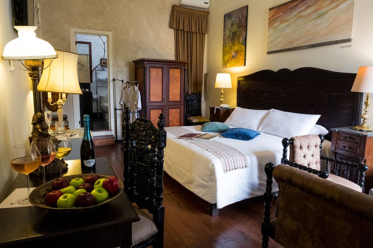 Ein Hotelzimmer mit einem Bett, zwei Stühlen, einem Tisch mit Obstschale und Weingläsern sowie einem Schrank.