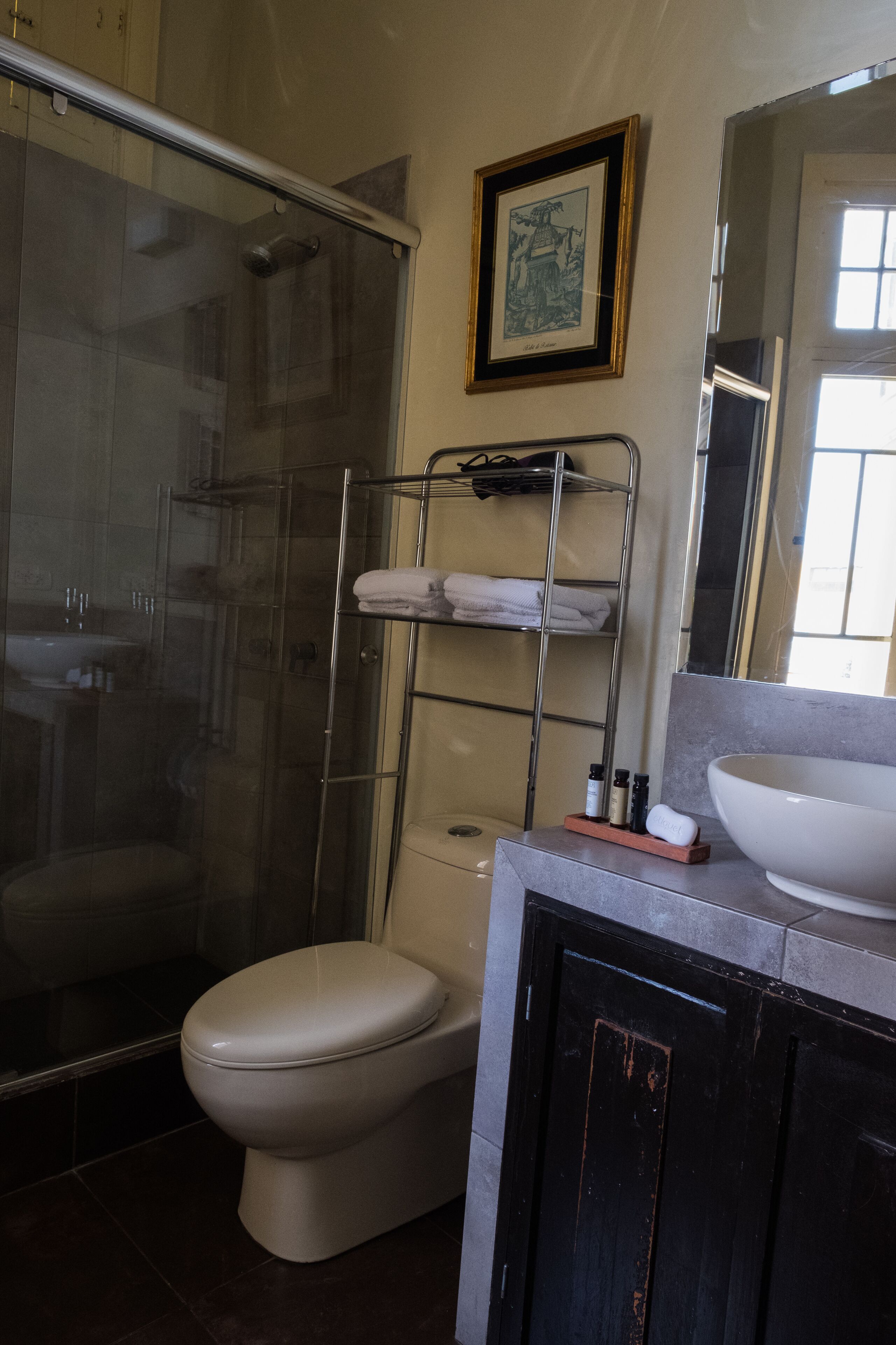 A bathroom with a glass-enclosed shower, a toilet, a sink, and a shelf with towels and toiletries.