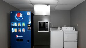 Habitación con una máquina expendedora azul llena de diversas bebidas, un refrigerador y una lavadora.