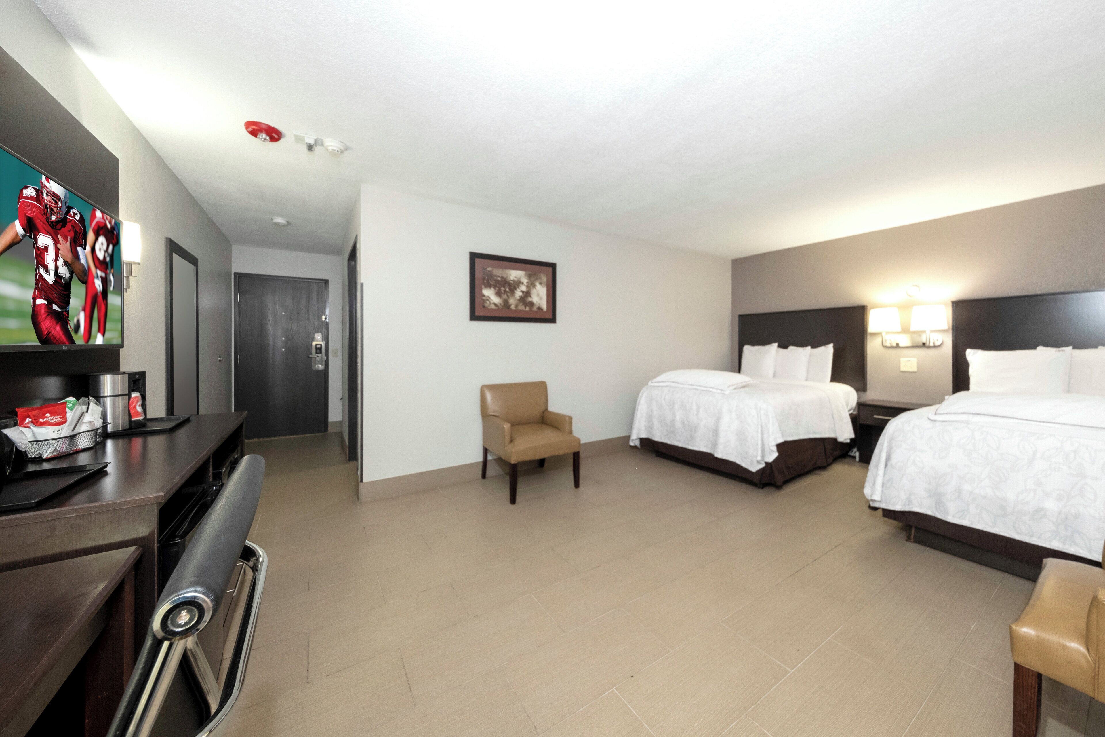 Habitación de hotel con dos camas, un escritorio con silla, un televisor y un cuadro en la pared.