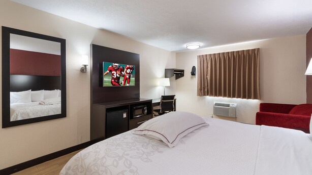 A hotel room with a large bed, a flat-screen TV mounted on the wall, a desk with a chair, a red armchair, and a window with curtains.