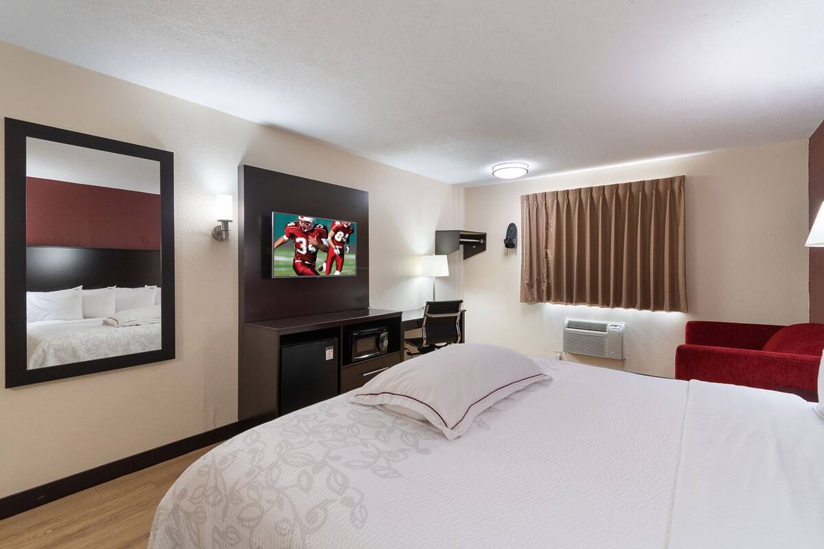 A hotel room with a large bed, a flat-screen TV mounted on the wall, a desk with a chair, a red armchair, and a window with curtains.