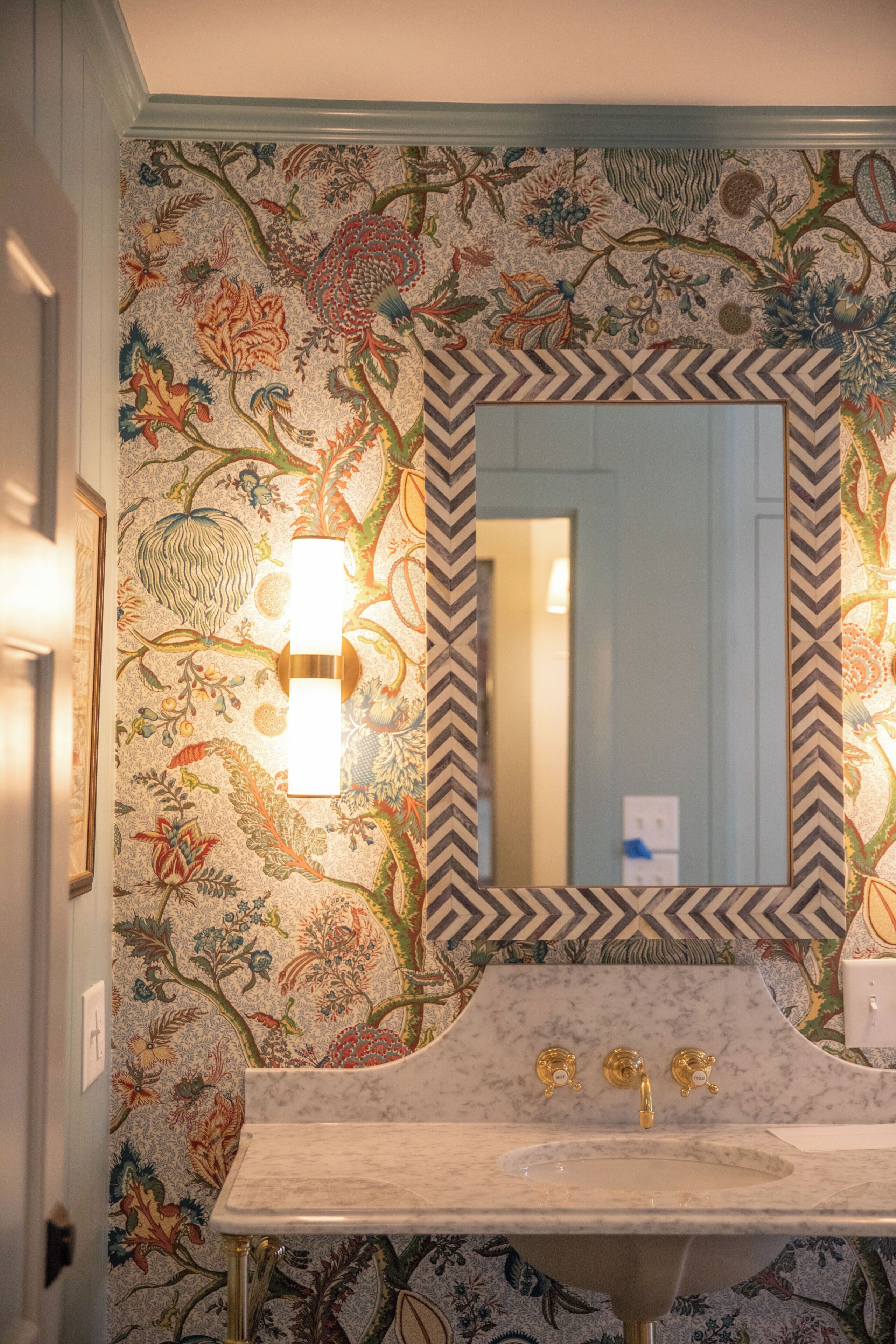 A bathroom with a floral wallpaper, a mirror with a chevron frame, and a marble sink with gold fixtures.